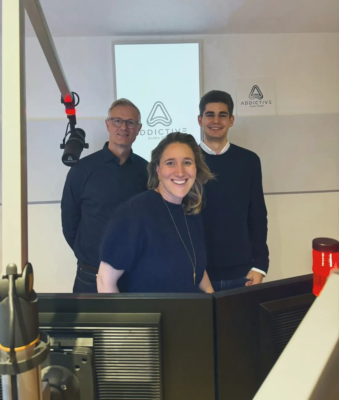 Drei Personen im Studio von Addictive Audio, im Hintergrund das Firmenlogo