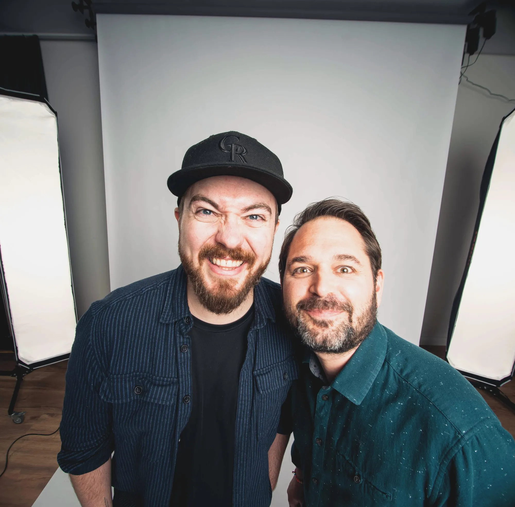 Christian Grollmann mit Kappe und Martin Putz lachen gemeinsam in die Kamera, hinter ihnen ein Fotohintergrund und zwei Softboxen.