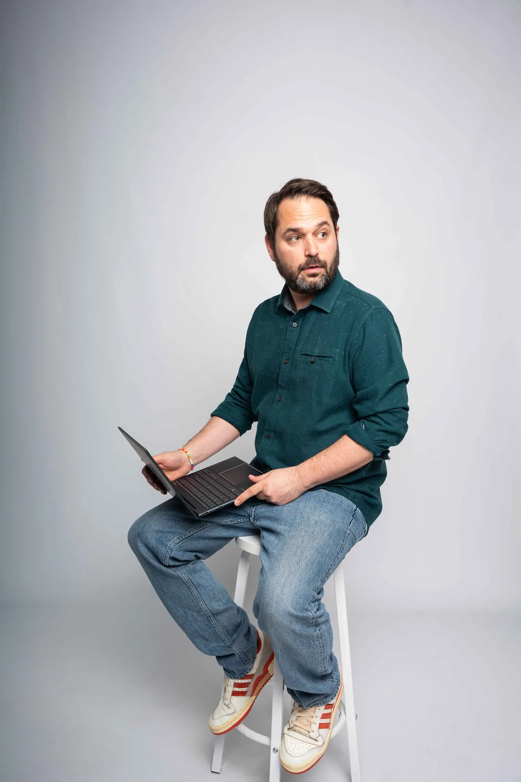 Christian Grollmann sitzt mit einem Laptop in der Hand auf einem Hocker und schaut nachdenklich zur Seite.