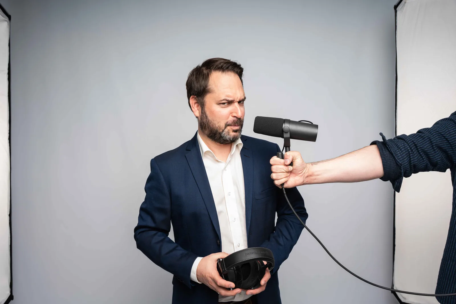 Christian Grollmann im Anzug hält ein VR-Headset, während ihm jemand mit ausgestrecktem Arm ein Podcast-Mikrofon ins Gesicht streckt, eine spielerische Szene aus dem Studio.