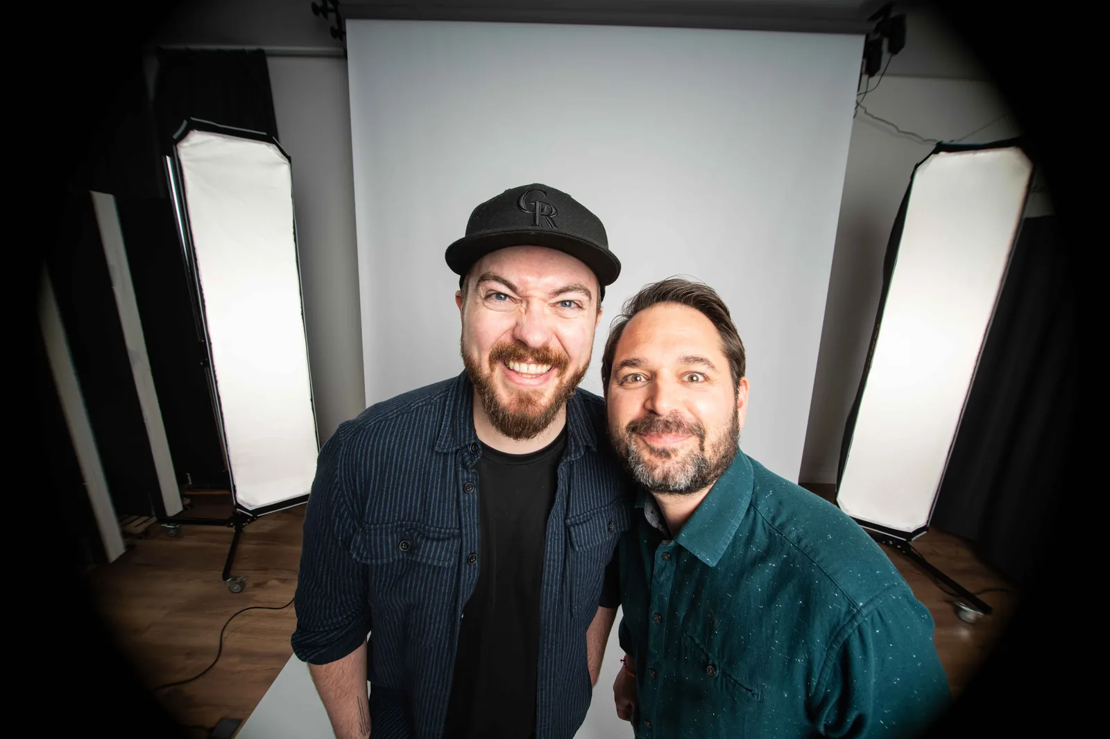 Christian Grollmann und Martin Putz lachen gemeinsam ins Fischaugen-Objektiv im Aufnahmestudio, umgeben von Softboxen und Studio-Equipment.